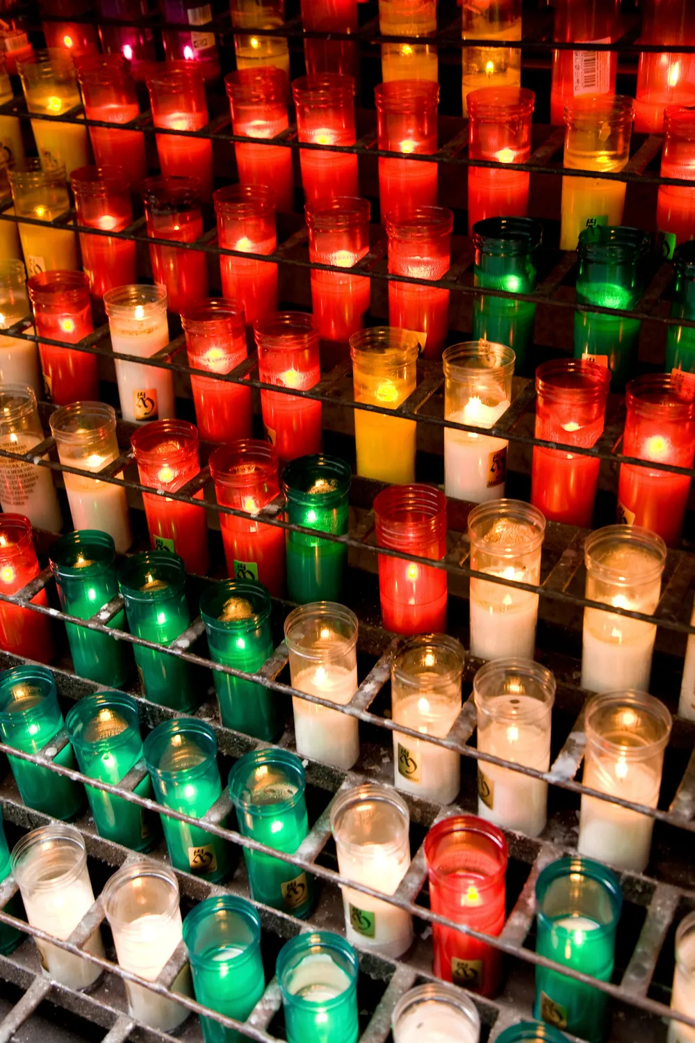 Fileiras de velas votivas acesas em copos de vidro colorido, em tons de vermelho, verde e dourado